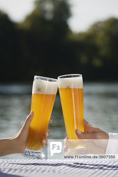 Junges Paar prosten sich mit Weizenbier zu Biergarten am Seehaus Englischer Garten Nahaufnahme der Gläser und Hände