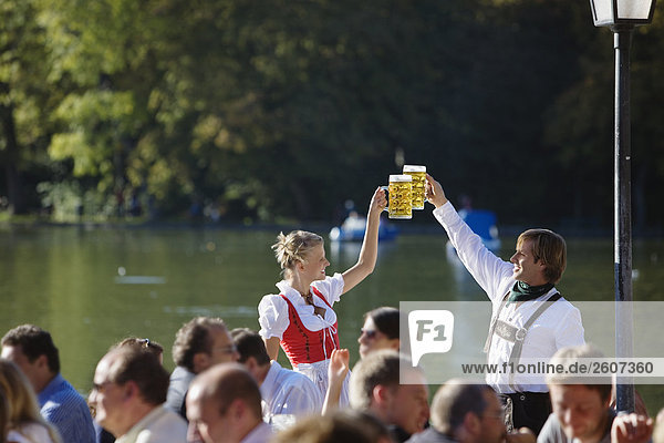 Junges Paar in Tracht steht im Biergarten am Seehaus Englischer Garten prosten sich mit einer Mass Bier zu Menschenmenge im Vordergrund