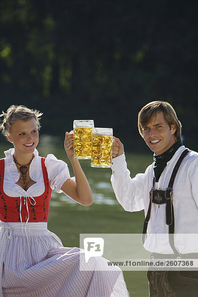Junges Paar in Tracht steht vor See prosten sich mit einer Mass Bier zu Biergarten am Seehaus Englischer Garten