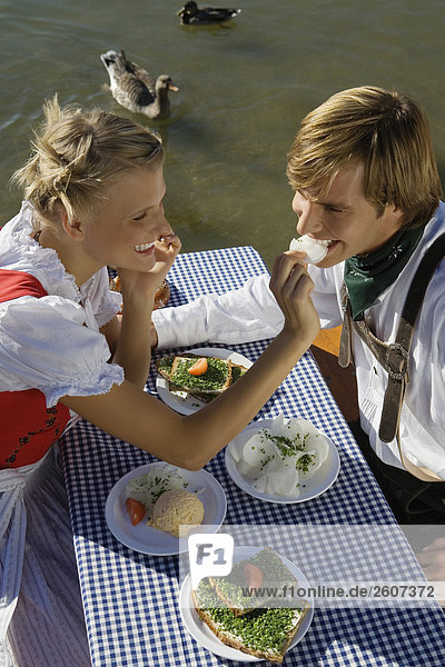 Junges Paar in Tracht sitzt am Biertisch mit Schnittlauchbrote Radi Obatzda und Breze Frau füttert Mann Biergarten am Seehaus Englischer Garten