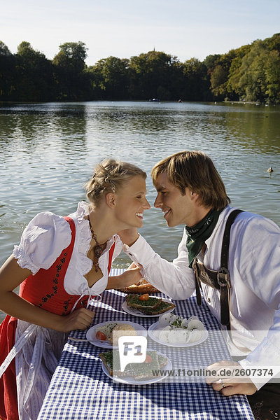 Junges Paar in Tracht sitzt am Biertisch mit Schnittlauchbrote Radi Obatzda und Breze Mann streichelt Frau Biergarten am Seehaus Englischer Garten