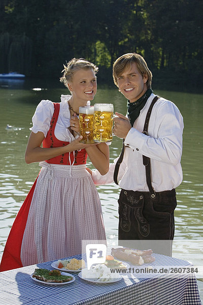 Junges Paar in Tracht steht am Biertisch mit essen prosten sich mit einer Mass Bier zu im Biergarten am Seehaus Englischer Garten