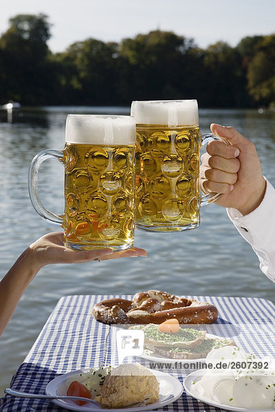Junges Paar sitzt am Biertisch mit Essen halten Mass Bier in der Hand Biergarten am Seehaus Englischer Garten Nahaufnahme der Masskrüge