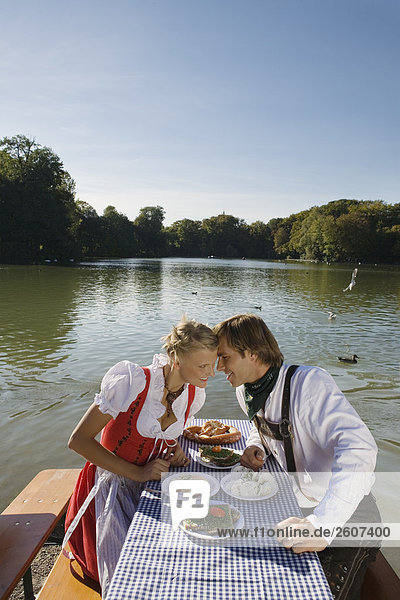 Junges Paar in Tracht sitzt am Biertisch mit Schnittlauchbrote Radi Obatzda und Breze Stirn an Stirn Biergarten am Seehaus Englischer Garten