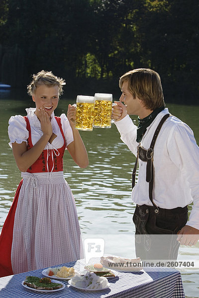 Junges Paar in Tracht steht am Biertisch mit Essen prosten sich mit einer Mass Bier zu Biergarten am Seehaus Englischer Garten