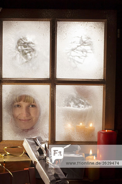Österreich  Salzburger Land  Junge Frau schaut durchs Fenster  Weihnachtspäckchen und Kerzen im Vordergrund