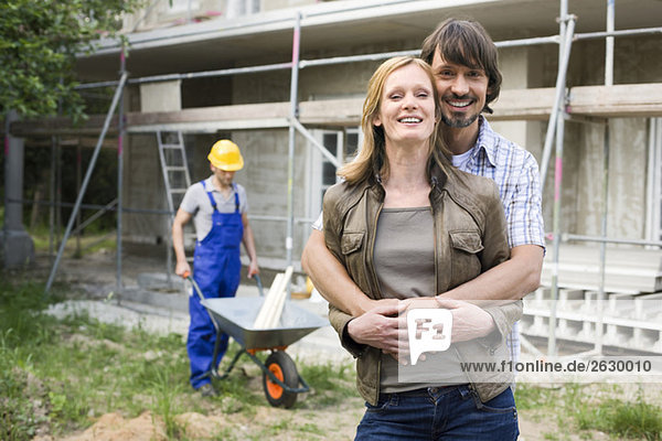 Young couple at site embracing  construction worker in background