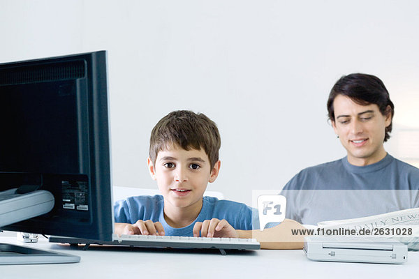 Kleiner Junge am Computer  lächelt in die Kamera  Vater liest Zeitung im Hintergrund
