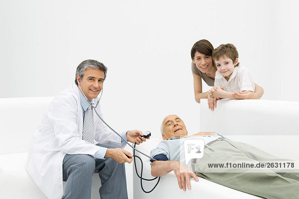 Arzt  der den Blutdruck eines älteren Mannes misst  Familie  die sich auf die Couch lehnt  alle lächeln in die Kamera.