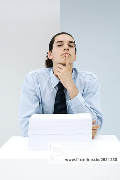 Young office worker behind stack of papers  hand under chin  looking at camera
