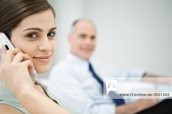 Professional woman using cell phone  smiling at camera  male colleague in background