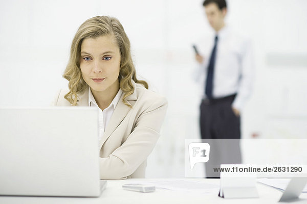 Professionelle Frau mit Laptop am Schreibtisch  Mann steht im Hintergrund