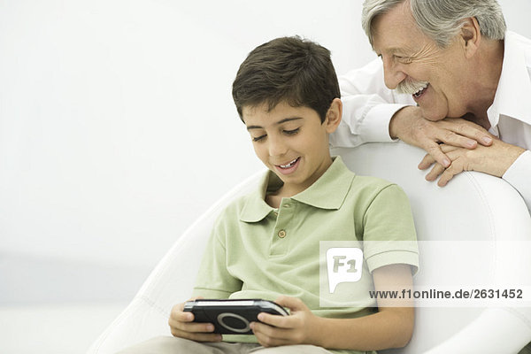 Grandson playing handheld video game  grandfather watching over shoulder