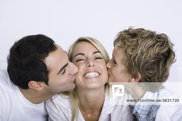 Lächelnde Frau  die von ihrem Mann und ihrem kleinen Sohn auf jede Wange geküsst wird.