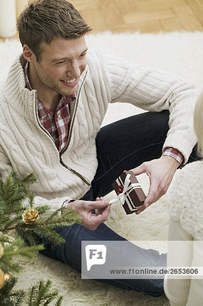 Paar neben Christbaum Mann öffnet Weihnachtspäckchen
