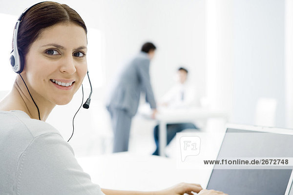 Frau trägt ein Headset  arbeitet am Schreibtisch  lächelt über die Schulter vor der Kamera