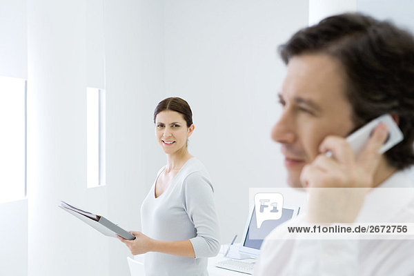 Professional woman in office  smiling at camera  male colleague using cell phone in foreground