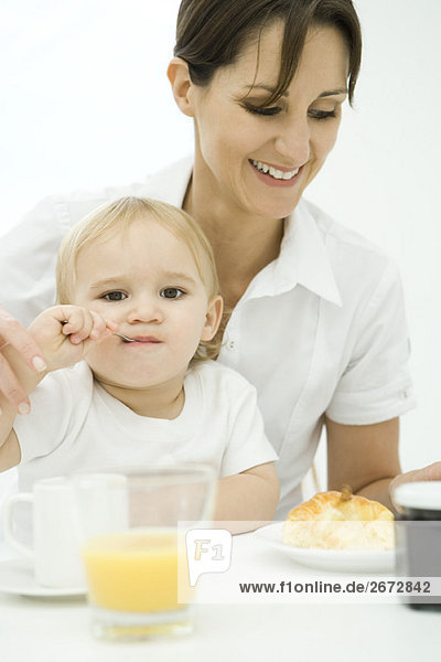 Professionelle Frau sitzt am Frühstückstisch  Kleinkind sitzt auf dem Schoß  Löffel im Mund