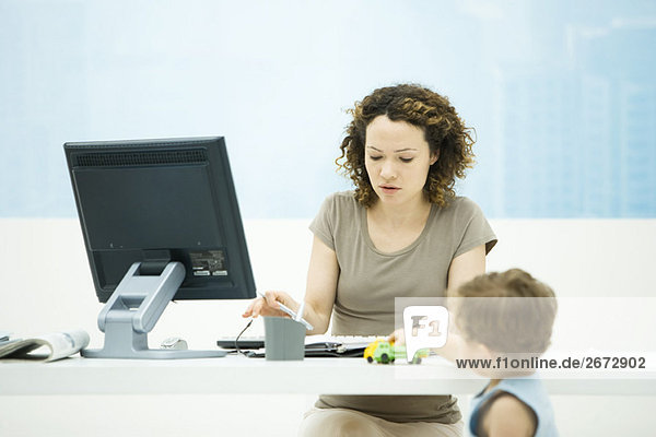 Frau tippt im Büro  Kleinkind-Sohn spielt mit Spielzeug-LKW auf Schreibtisch