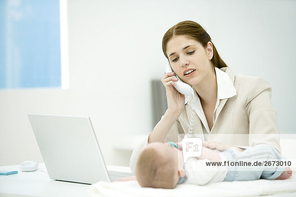 Professionelle Frau berührt Kleinkind auf dem Schreibtisch liegend  telefonierend