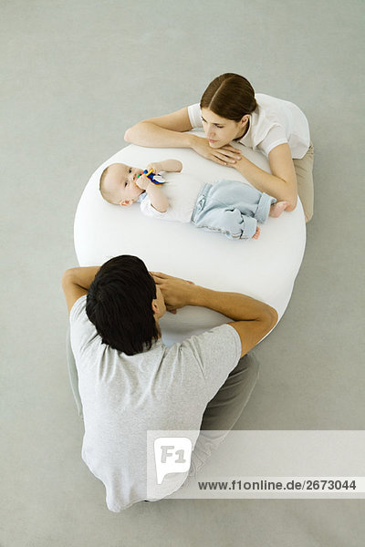 Junge Eltern  die sich gegen den Hocker lehnen und das zwischen ihnen liegende Baby anschauen