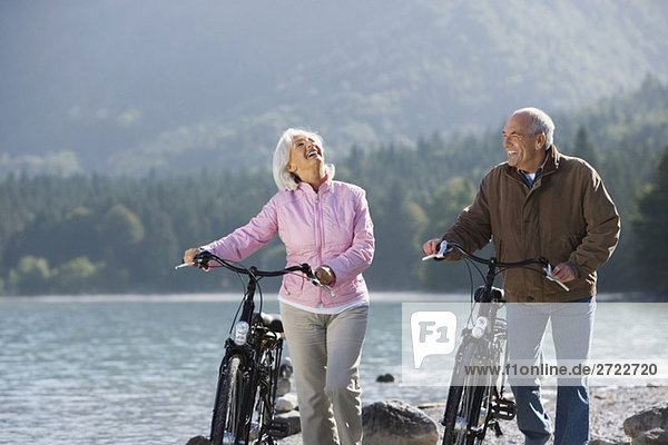 Germany  Bavaria  Walchensee  Senior couple pushing bikes across lakeshore