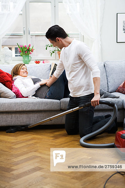 A pregnant woman lying on a couch and a man vacuuming Sweden.