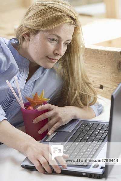 Blonde Frau mit Laptop und Cocktail auf dem Bett
