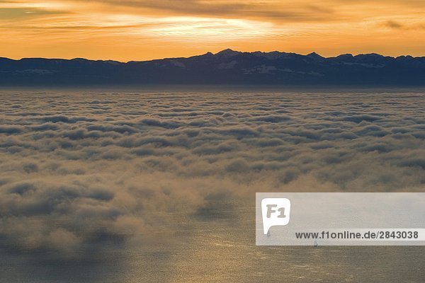 View from mount cypress mountain with Sailboats appearing in ocean covered with fog and clouds with sun setting behing mountains in west vancouver british columbia canada.
