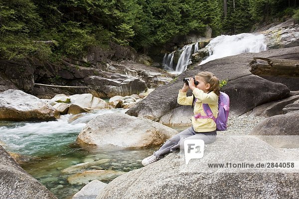 Young girl sitting on a rock and looking through binoculars in front of waterfalls in Golden Ears Provincial Park in Maple Ridge  British Columbia  Canada