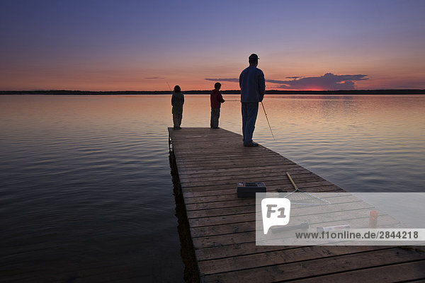 A father with his two sons fishing at the end of a wharf on Lake Audy at sunset  Riding Mountain National Park  Manitoba  Canada
