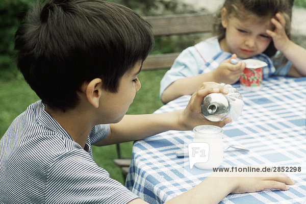 Junge sitzt mit Schwester am Tisch und gießt Zuckerjoghurt.