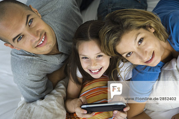 Mädchen liegt zwischen den Eltern  spielt Handheld-Videospiel  alle lächeln vor der Kamera