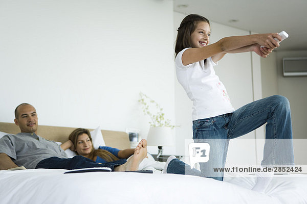 Mädchen im Bett spielt Videospiel mit drahtlosem Controller  Eltern sehen im Hintergrund zu.