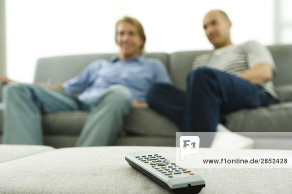 Zwei Männer sitzen auf dem Sofa und konzentrieren sich auf die Fernbedienung im Vordergrund.