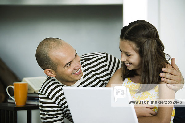 Vater und Tochter sitzen mit Laptop und lächeln sich an.