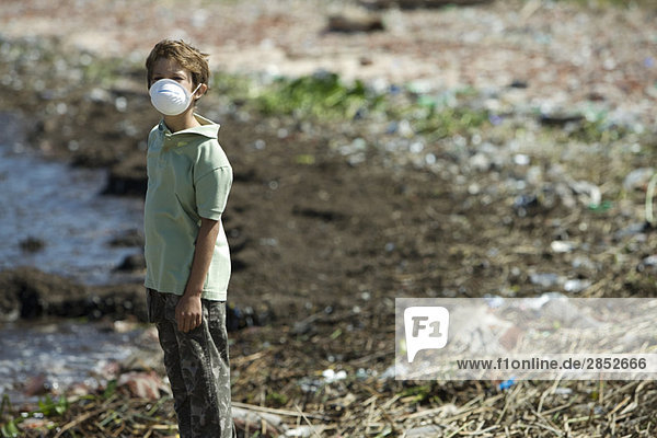 Junge steht am verschmutzten Ufer und trägt eine Verschmutzungsmaske.