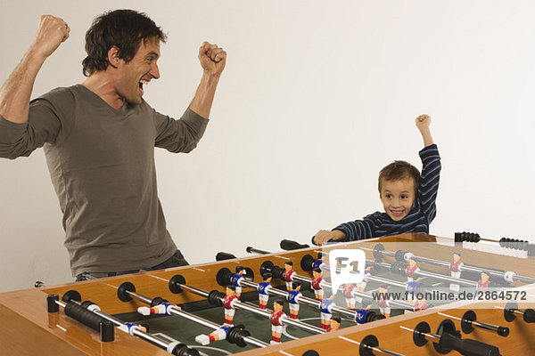 Vater und Sohn (4-5) spielen Tischfußball und jubeln zusammen.