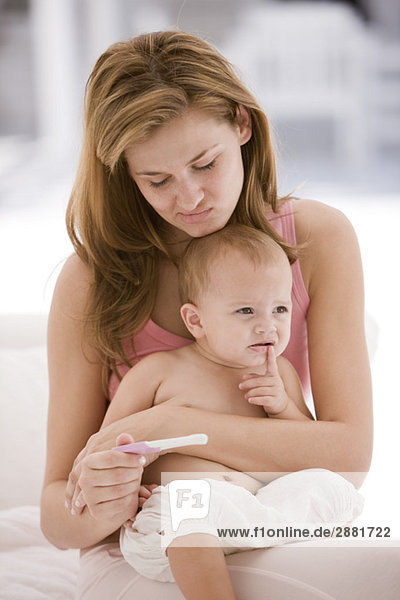 Frau betrachtet einen Schwangerschaftsteststab mit einer Tochter  die auf ihrem Schoß sitzt.