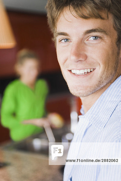 Porträt eines lächelnden Mannes mit einer Frau  die im Hintergrund Gemüse wäscht.