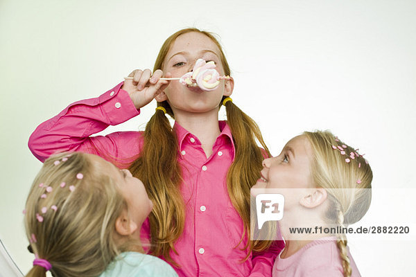 Ein Mädchen isst einen Lolli  während ihre beiden Freunde sie ansehen.