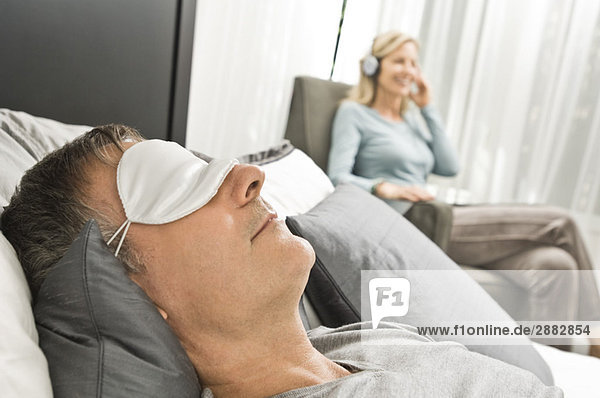 Man wearing an eye mask and his wife listening to music in the background