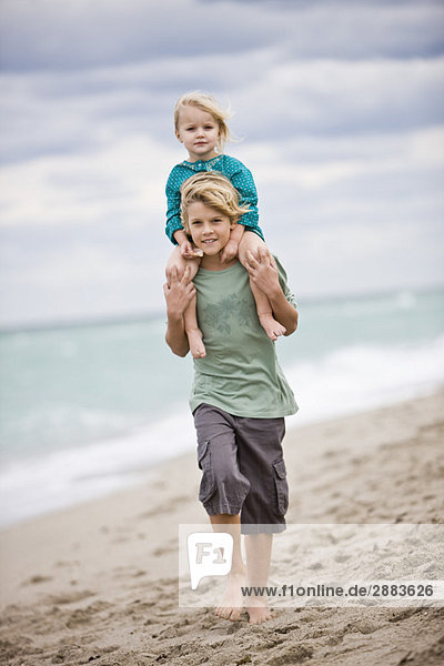 Junge der seine Schwester auf den Schultern am Strand trägt.