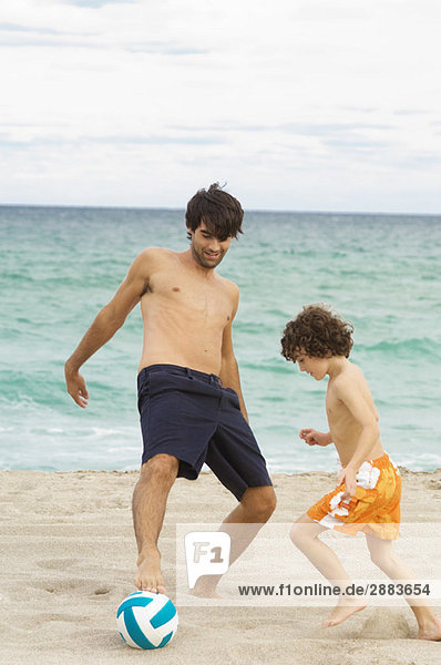 Junge spielt Fußball mit seinem Vater am Strand.