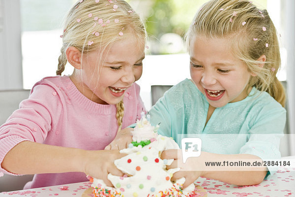 Zwei Mädchen  die spielerisch ihre Hände in einen Geburtstagskuchen stecken.