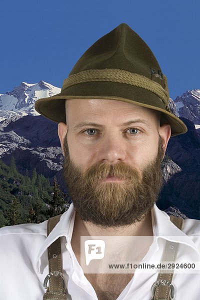 A man wearing an alpine hat and lederhosen with a mountain backdrop