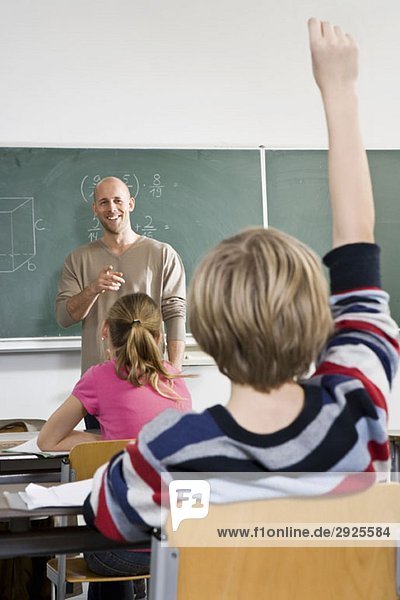 Rückansicht eines Jungen  der seine Hand in einem Klassenzimmer hebt.