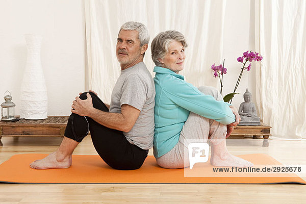 A senior man and a senior woman sitting back to back on an exercise mat