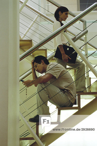 Mann mit der Hand auf dem Kopf sitzend auf der Treppe mit Blick auf das Handy  Frau  die vorbeigeht