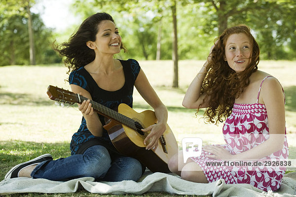 Two young women sitting on blanket outdoors  one playing acoustic guitar
